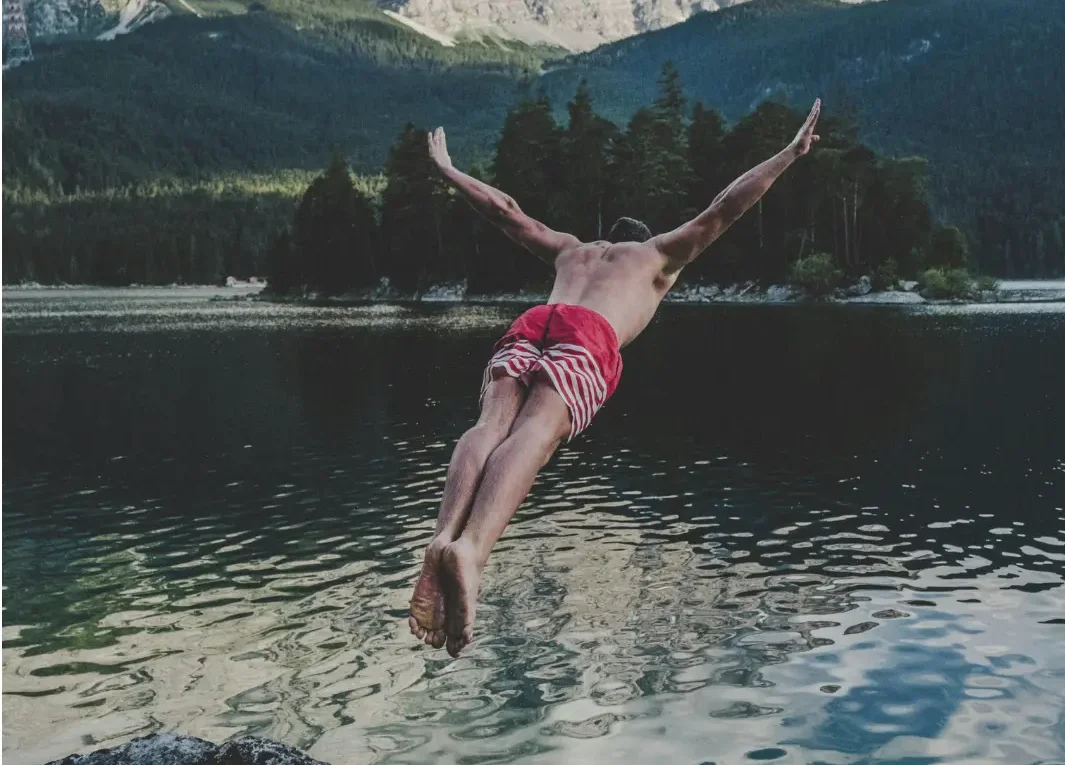 Man diving from cliff into blue ocean with mountains in the background